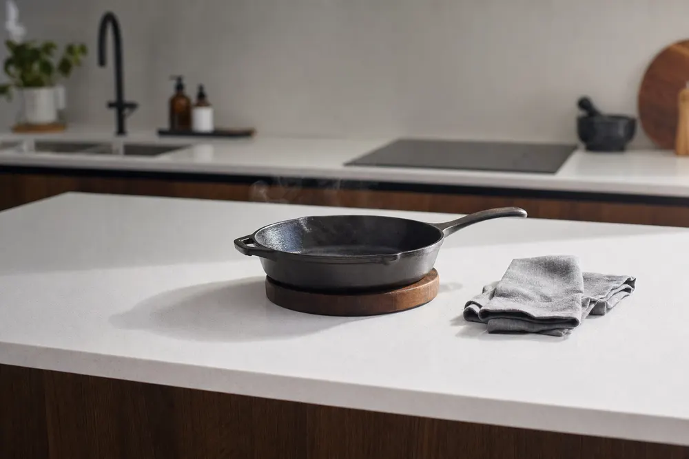 A cast iron pan resting on a timber heat mat on a white speckled engineered stone benchtop